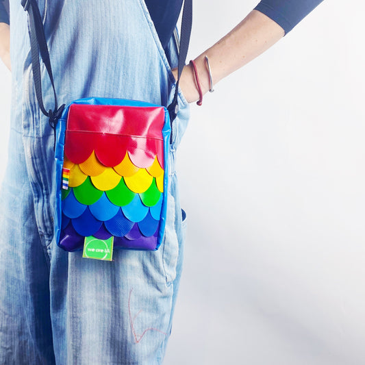 A person wearing blue denim overalls is holding a colorful, rainbow-mermaid-patterned mini crossbody bag with a zipper and an adjustable strap.
