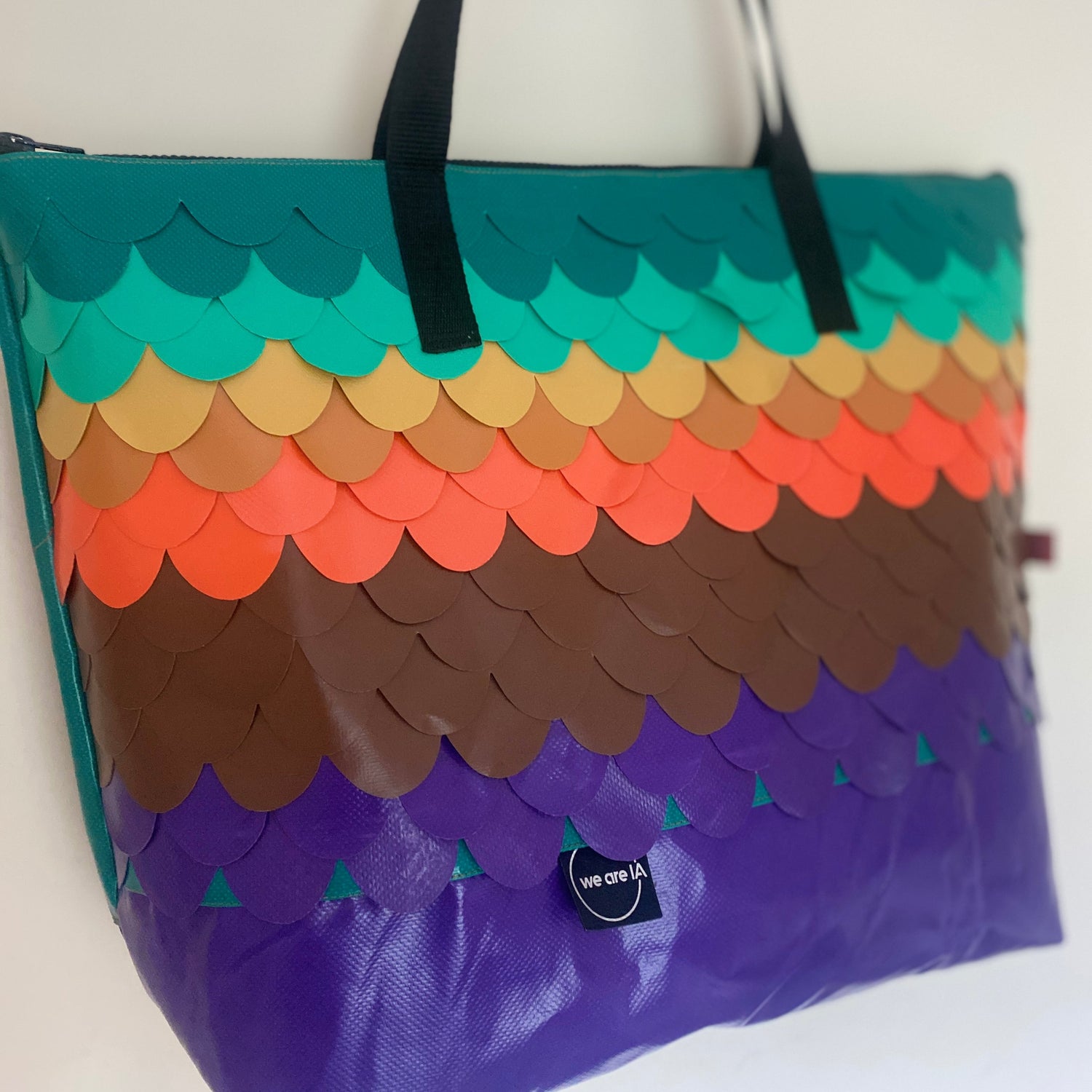 A large tote bag with a colorful scalloped scale pattern in teal, green, tan, orange, brown, and purple, featuring black shoulder straps and a small black brand tag that reads 'we are IA'