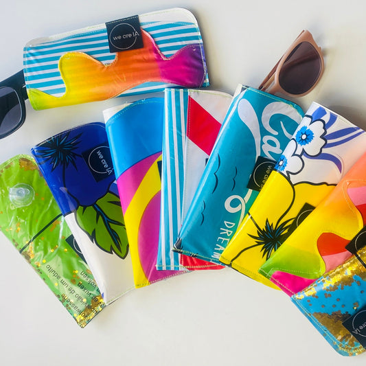 A group of colourful sunglasses cases made from recycled inflatables and camping mattresses, arranged in a fan shape on a white background, with several pairs of sunglasses resting on top. The cases feature vibrant tropical and abstract patterns in blue, yellow, pink, green, and white.